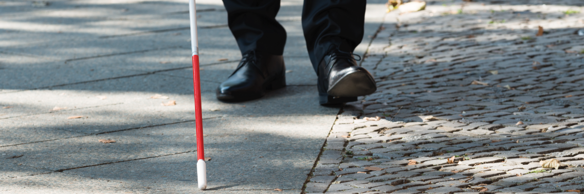 A white cane scans a sidewalk. In the background, men's shoes trail behind the tip of the cane.