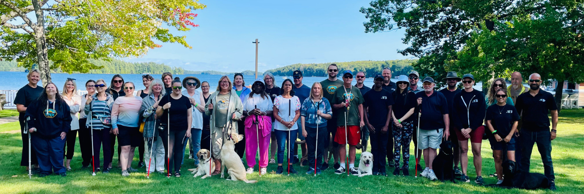 Une photo de groupe prise au bord de l’eau au Centre Lake Joe d’INCA. Le groupe est composé de membres de l’équipe de Réadaptation en déficience visuelle, de l’équipe des Chiens-guides d’INCA et de participants au Programme d’immersion en orientation et mobilité. 