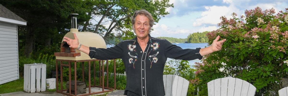 Jim Cuddy is standing in black shirt and jeans at the campfire circle in front of white Muskoka chairs and the outdoor pizza oven. He has his arms outstretched and is smiling.