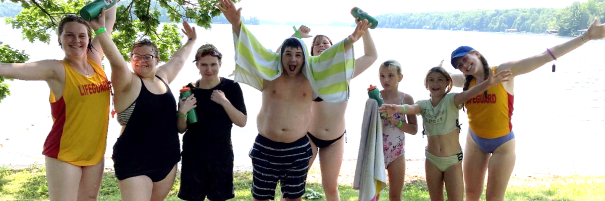 Six children and youth stand at the water’s edge wearing their bathing suits, smiling with their arms outstretched. Two lifeguards stand on opposite sides.