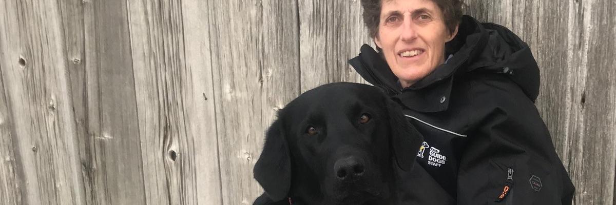  Outdoors, Dr. Bennett stands against a wooden fence and has around a black lab future guide dog. 