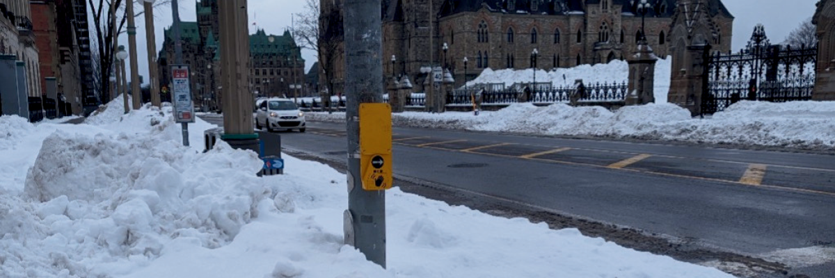 An APS button fixed to a pole at a crosswalk in Ottawa, with Parliament buildings in the background. The pole is surrounded by a large snowbank that reaches almost as high as the button and extends approximately a metre outward, making it impossible for pedestrians to reach it.