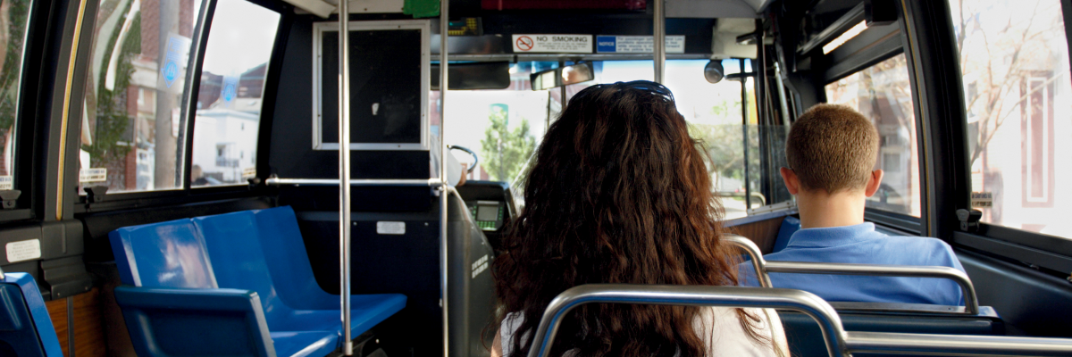 The interior of a city bus. The view is from the rear of the bus, looking towards the front. A few passengers are seated.