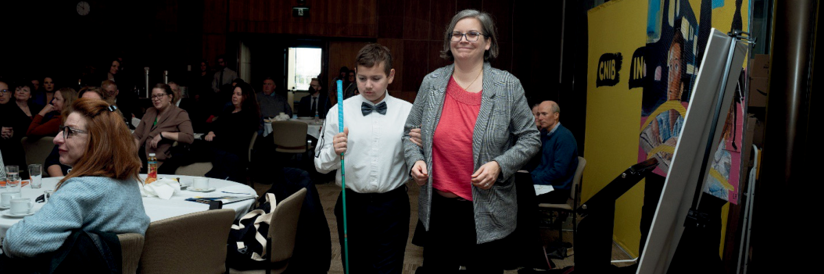 Dawn Pickering sourit en se dirigeant avec son fils, Ollie, vers la scène pour le panel des familles lors du lancement parlementaire de la Charte des droits des enfants d’INCA. On voit les participants devant une grande bannière d'INCA à l'arrière-plan. Ollie porte une chemise blanche avec un nœud papillon noir et tient sa canne blanche.