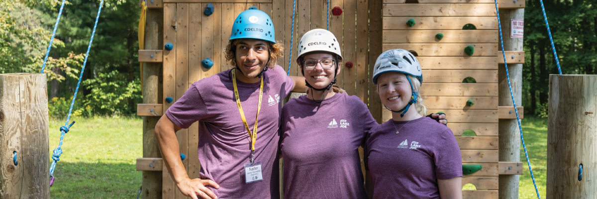 Three young CNIB Lake Joe staff members stand in front of the new climbing tower at Lake Joe. They are wearing helmets and CNIB Lake Joe staff shirts.