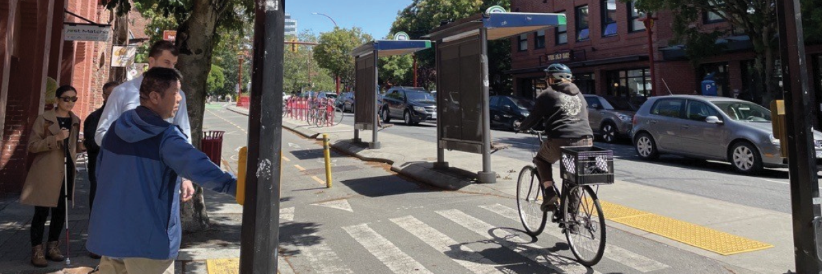 Un piéton et son chien guide tentent de traverser la rue pour se rendre à un arrêt d’autobus sur îlot. Le piéton appuie sur un bouton de passage pour piétons tandis qu'un cycliste traverse à vélo le passage pour piétons. 