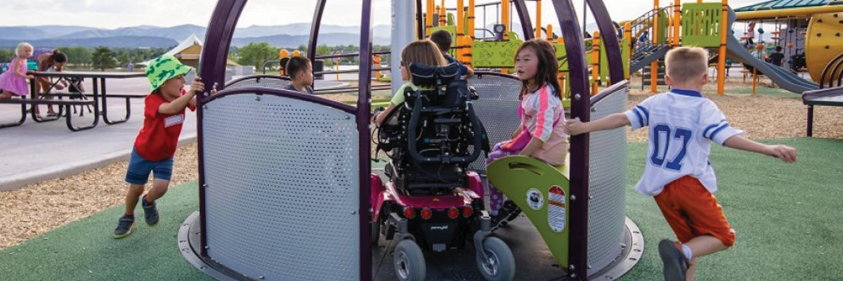 Des enfants jouent sur une aire de jeux accessible par une journée d'été ensoleillée. Un enfant en fauteuil roulant se trouve sur un carrousel accessible aux fauteuils roulants avec trois autres enfants. Deux autres enfants courent et tirent le carrousel pour le faire tourner pour les enfants assis dessus. 