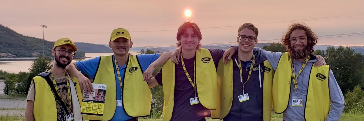 Cinq collecteurs de fonds travaillant pour le compte d'INCA se tiennent côte à côte, les bras enlacés, devant un coucher de soleil d'un rouge éclatant. Ils portent tous un gilet jaune d'INCA et un cordon avec photo d'identité. Les deux collecteurs de fonds de gauche portent également une casquette de baseball jaune d'INCA et tiennent de grandes cartes présentant des faits sur la cécité.