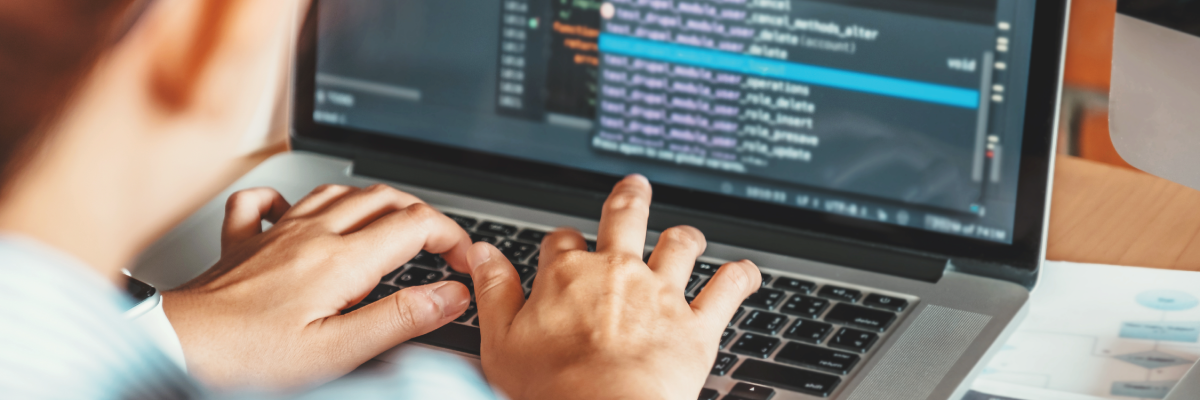 Une personne tape sur un ordinateur portable. La photo est prise par-dessus son épaule et l'écran de son ordinateur portable affiche des lignes de code, ce qui suggère qu'elle est en train de programmer ou de coder.