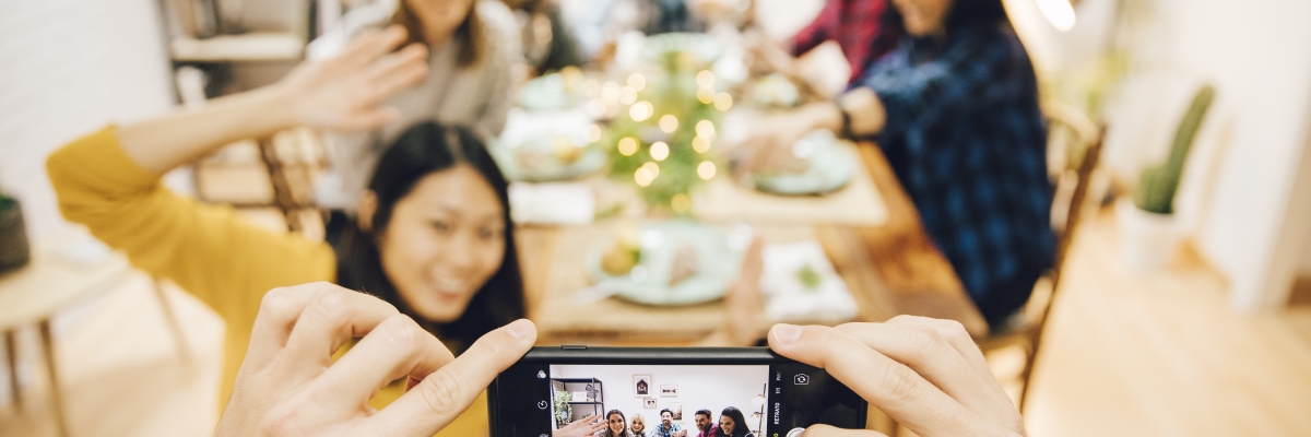 une personne prend une photo de famille à l'aide d'un iPhone. La famille est assise autour d'une table dans une salle à manger décorée de lumières festives.  