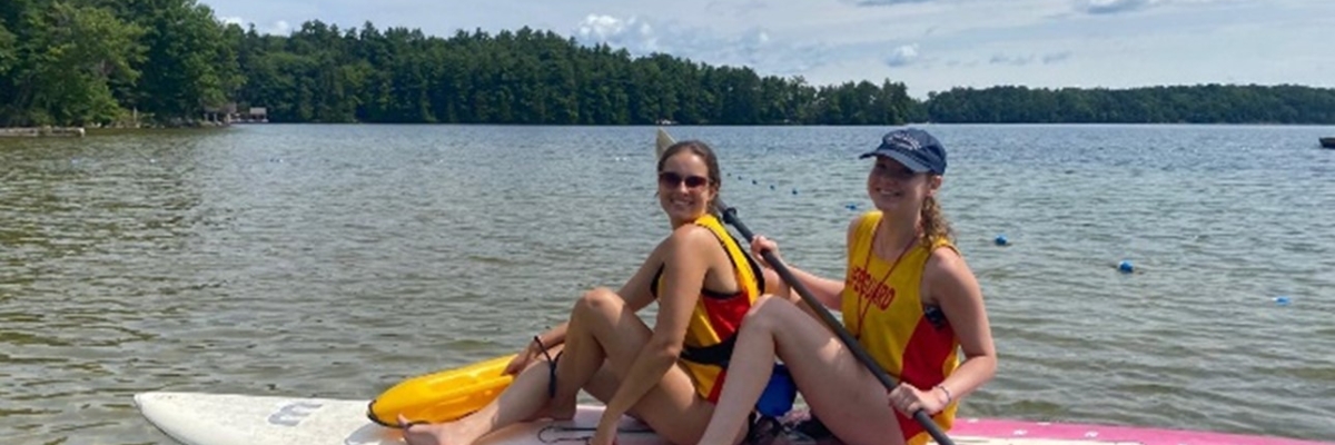 Deux jeunes femmes portant des maillots de surveillantes de baignade sont assises sur une planche à pagaie sur le lac. 