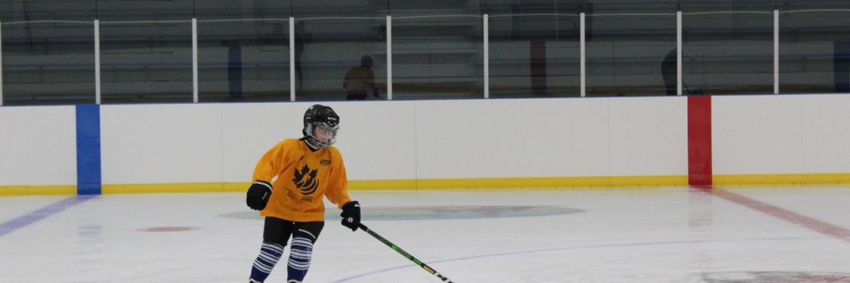 Un jeune garçon patine sur la glace d’une patinoire. Il porte un casque, un maillot de hockey jaune, un pantalon, des patins noirs et tient un bâton de hockey d'une main 