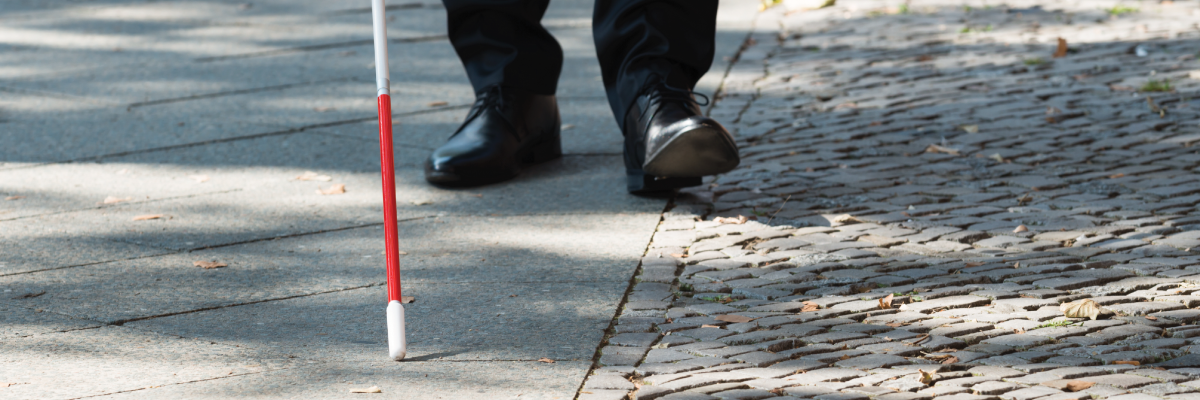 Une canne blanche balaie un trottoir. En arrière-plan, on aperçoit des chaussures d’un homme derrière la pointe de la canne.