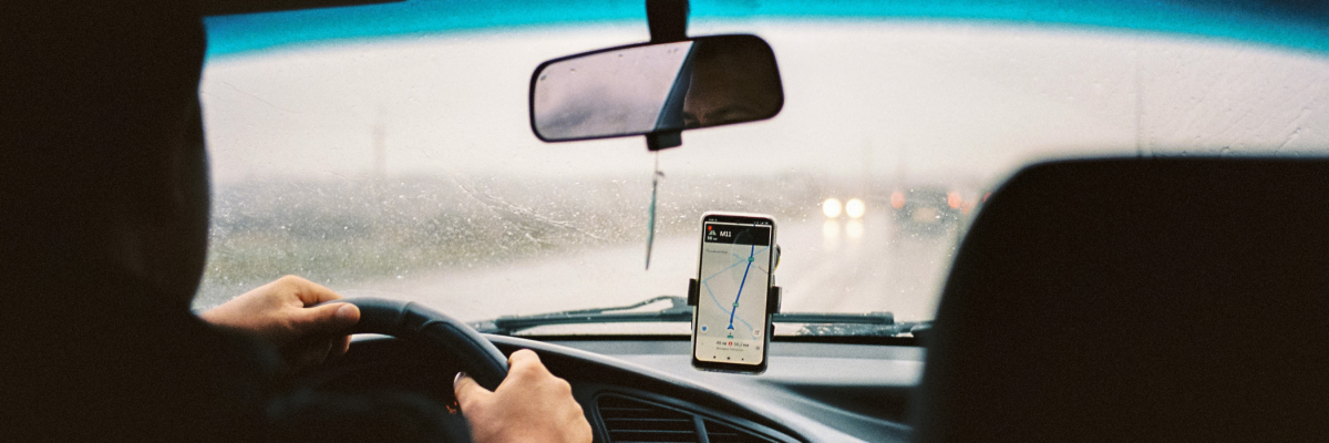 À l'intérieur d'une voiture de covoiturage, la photo est prise du point de vue du passager sur la banquette arrière. Il y a un téléphone intelligent monté sur le tableau de bord de la voiture affichant une application de rideshare. Le conducteur a les mains sur le volant et il pleut à l'extérieur. 