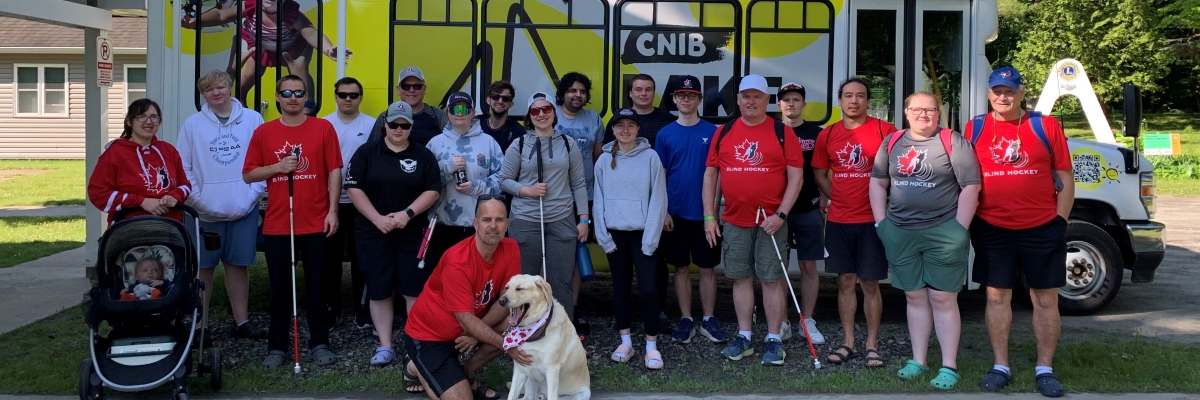Environ 20 personnes se tiennent devant le nouveau minibus du Camp Lake Joe d'INCA. Plusieurs portent des t-shirts rouges du Hockey sonore Canada et tiennent des cannes blanches. Un chien-guide se trouve devant.