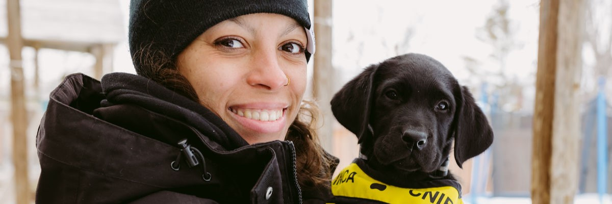 Une personne souriante, vêtue d'une veste d'hiver, tient dans ses bras un chiot noir portant un gilet du programme « Future Guide Dog » d'INCA.