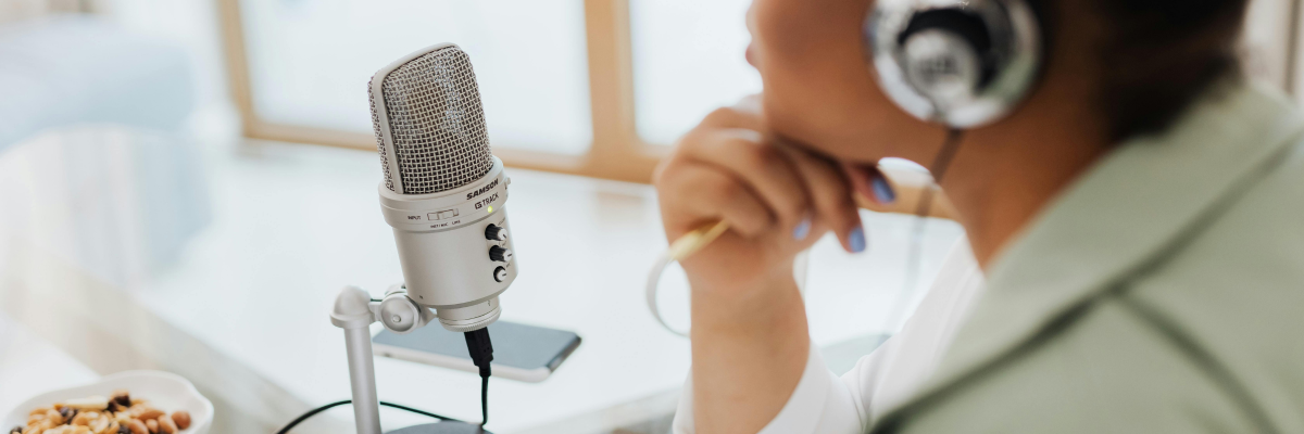 Une femme portant un casque audio est assise à un bureau, à côté d'un micro sur pied. Elle est en train d'enregistrer un podcast.