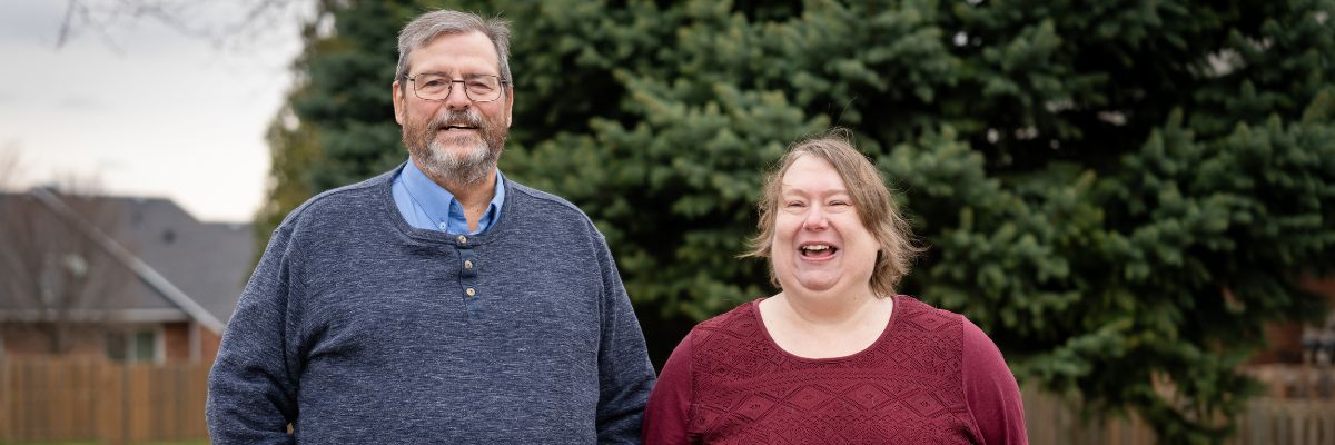 Un homme et une femme se tiennent côte à côte à l'extérieur, devant un grand conifère ; tous deux sourient et rient. L'homme, à gauche, a les cheveux gris, une barbe et porte des lunettes ; il est vêtu d'un pull Henley bleu marine par-dessus une chemise bleu clair. La femme, à droite, a les cheveux châtain clair et porte un haut texturé bordeaux foncé. L'image se présente sous la forme d'une grande bannière.