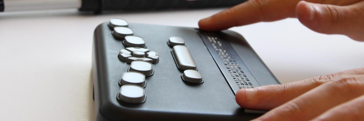 Hands reading the Orbit Braille Reader with a white cane in the background