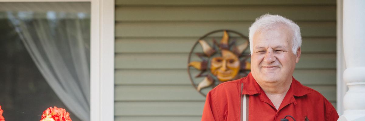 Man with grey hair, wearing a red shirt, holding his white cane by his side, standing on his front porch.