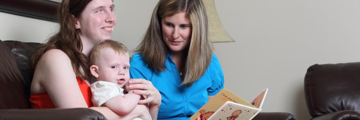 Two women sit on a couch. The woman on the left is holding an infant while the other woman is holding a storybook.