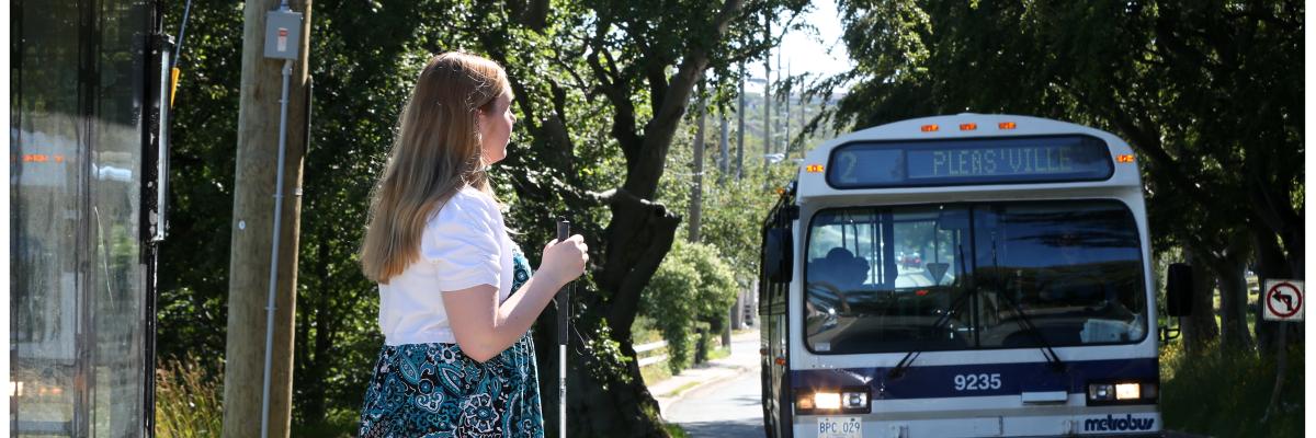 Young woman standing at a bus stop holding her white cane in front. The bus is approaching.