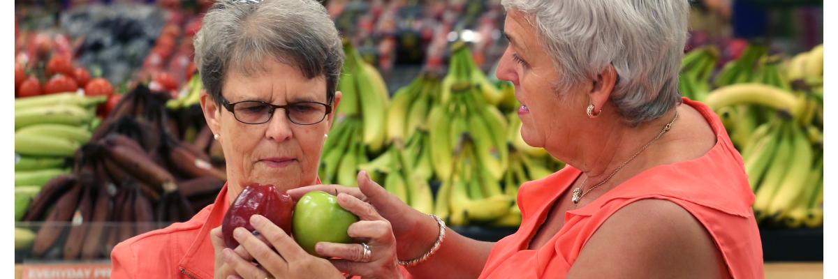 Two woman, wearing coral-coloured tops, are shopping for apples. 