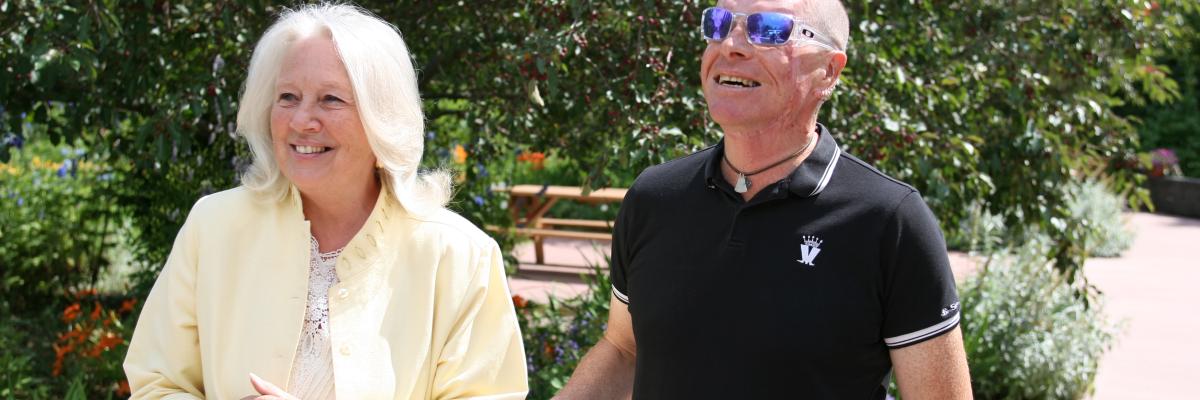 A woman in yellow guides a man in a black t-shirt. The man is holding on to her elbow. Both are smiling.