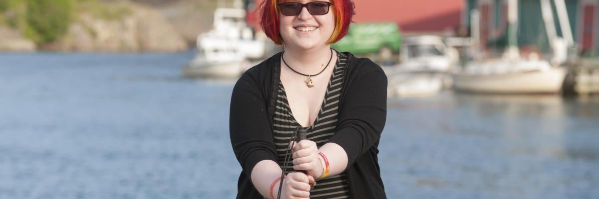 Young woman standing in front of a body of water with boats behind her, smiling at the camera holding her white cane in front of her.