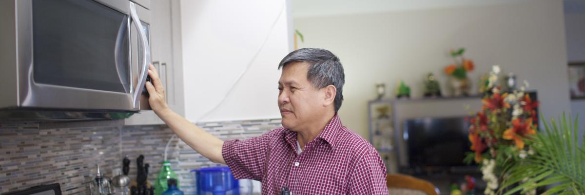 Man in a red plaid shirt is holding his white cane to his side while reaching for the microwave.