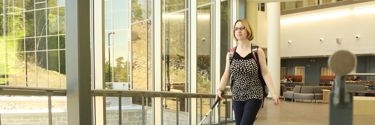 A young blond woman walking down a hallway with her white cane