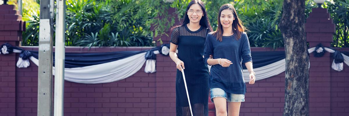 Deux femmes traversant la rue. Une utilise une canne blanche et l'autre la guide.