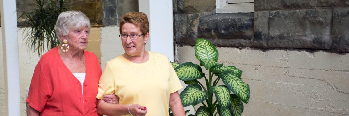 Elderly woman in red is walking and being guided by another woman who is wearing a yellow shirt and olive green pants.