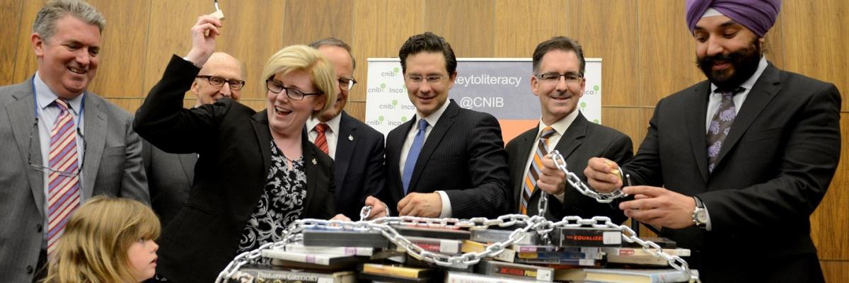 Minister Carla Qualtrough and Minister Navdeep Bains unlock a chain over top of books in a celebration of Canada's ratification of the Marrakesh Treaty. Members of Parliament Rob Oliphant, Pierre Poilievre and Brian Masse help unlock the books, while CNIB President and CEO John Rafferty and Craig Oliver overlook. 