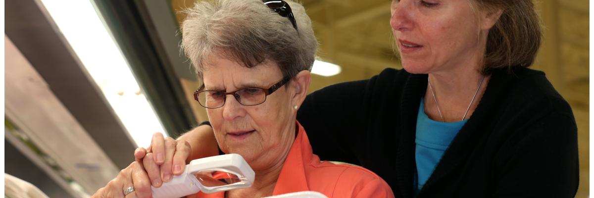 Older female holds a magnifier to a package. She is standing in front of another woman who is helping her with the magnifier.