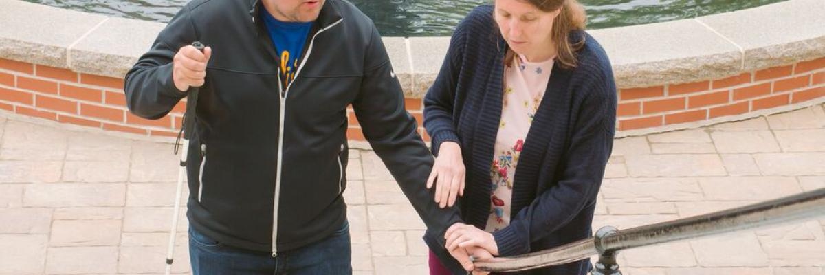 Young man starts walking up an outdoor staircase with his white cane. A young woman assists him to find the railing.