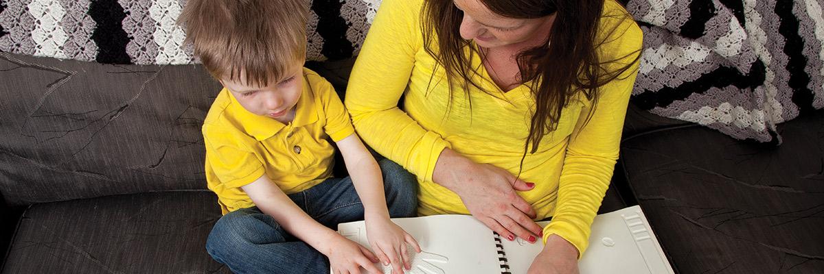 Un petit garçon assis à côté de sa mère sur un sofa lit un livre tactile.