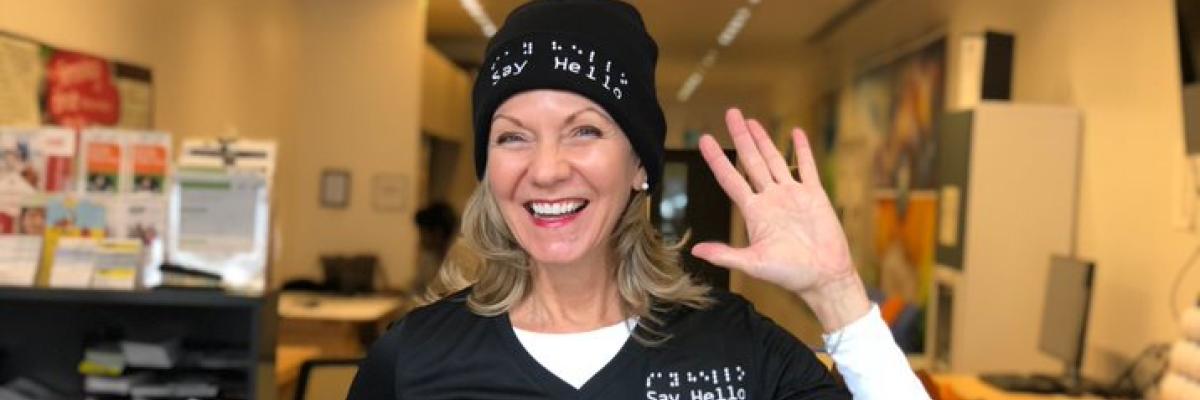 Say Hello 2 Blindness founder Denise Justin smiles and waves while wearing her company-branded shirt and toque.