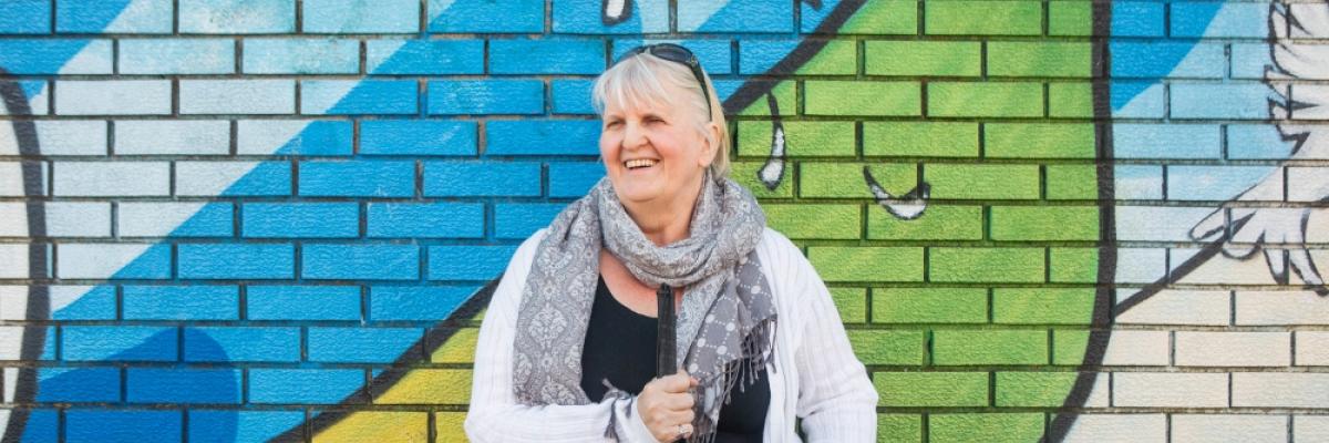 Wendy, holding her white cane, stands in front of a brick wall with a colourful mural.