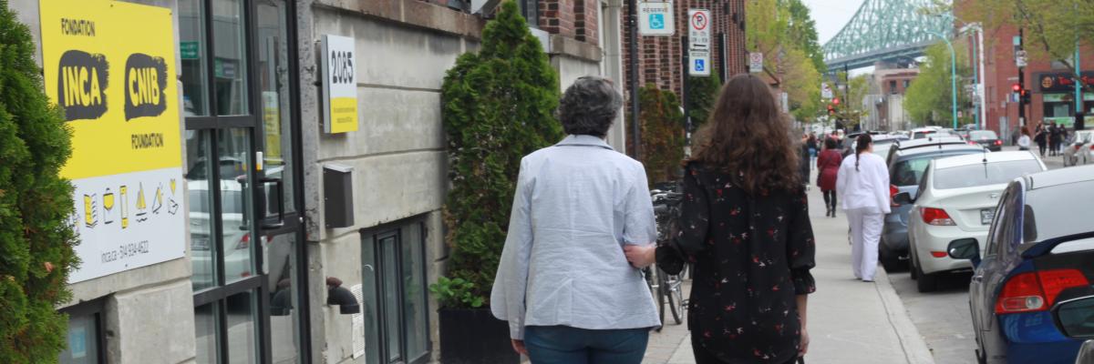 Une bénévole et une jeune femme aveugle marche en face du bureau d'INCA on voit le pont Jacques-Cartier à l'horizon