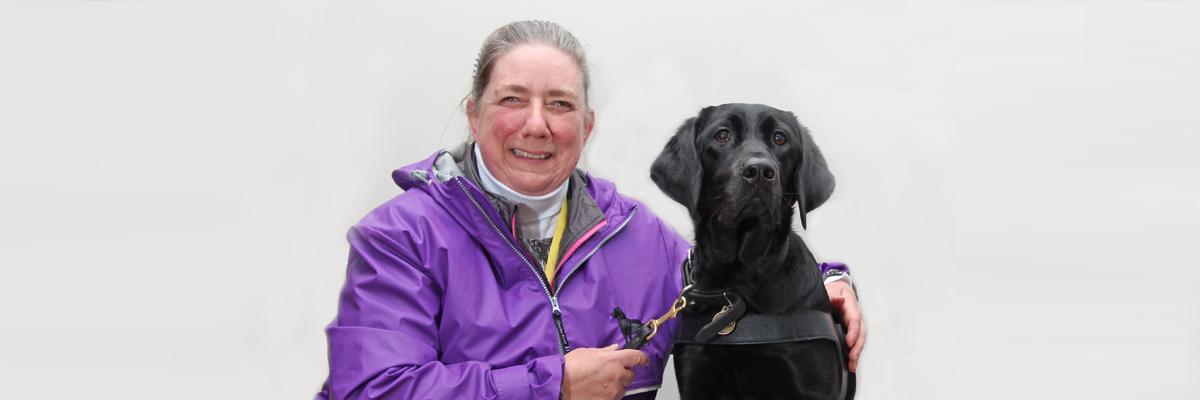 A woman in a purple jacket and her black Lab/Golden Retriever cross guide dog.