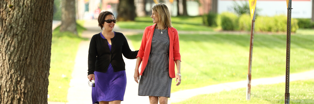 An example of the sighted guide technique. Two woman walk down a sidewalk together. A woman holds the guide's arm lightly above the elbow and allows the guide to walk one-half step ahead.