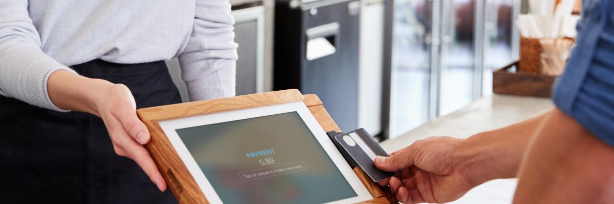 A person out of frame tapping their bank card on a touchscreen payment terminal, being presented by a café employee.