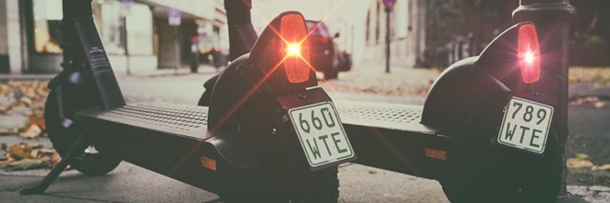  Two e-scooters sit side-by-side on a sidewalk. Their break lights are blinking red.