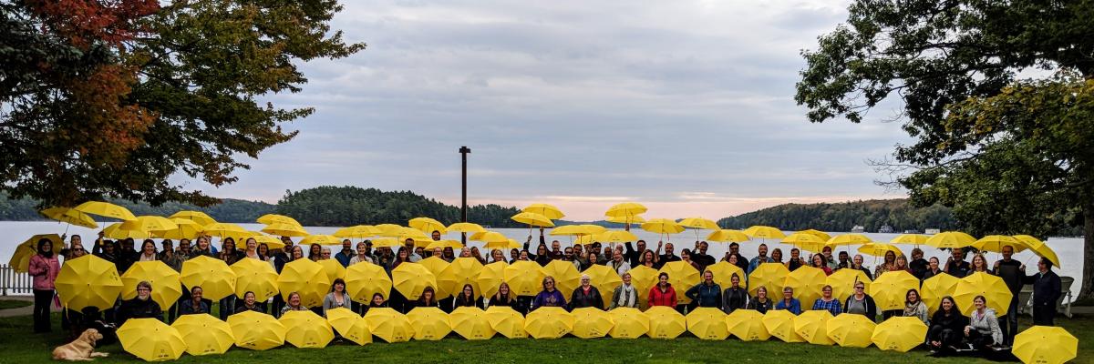 Un grand groupe de plus de 50 employés d’INCA tenant des parapluies jaunes d’INCA sur les rives du Centre Lake Joseph d’INCA.  