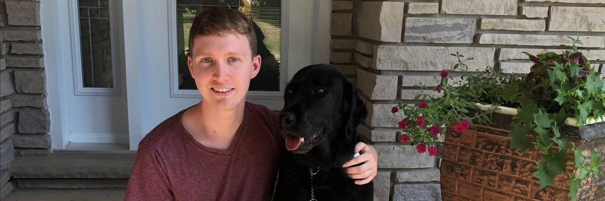 Jack McCormick sitting on the front step of house with his arm around his guide dog sitting at his side. 