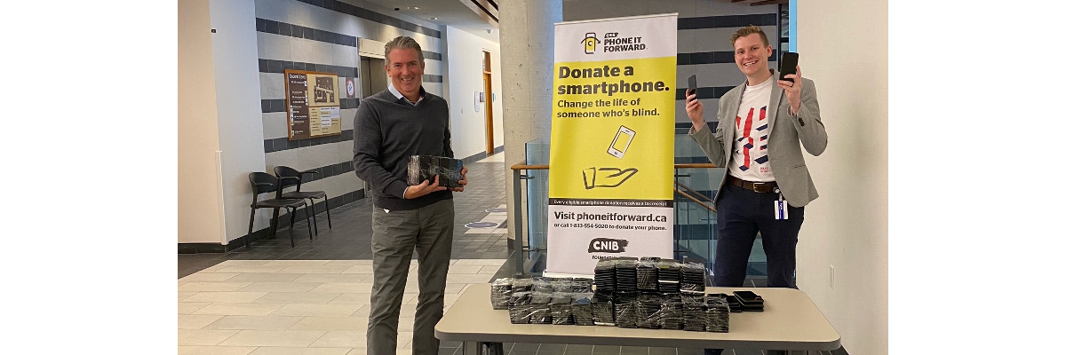 John M. Rafferty et Thomas Simpson, souriants, sont debout à une table et tiennent plusieurs téléphones intelligents donnés par la Banque du Canada. Sur la table sont également empilés des dons de téléphones intelligents. Derrière eux se trouve un présentoir portant la bannière du programme Rendre l'appareil.