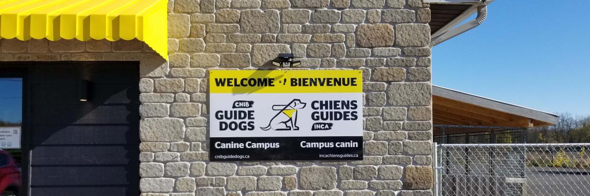 Une photo du panneau de bienvenue placé à l'extérieur du Campus canin. On peut y lire : « Welcome et Bienvenue » avec le logo des chiens-guides d'INCA au milieu du panneau
