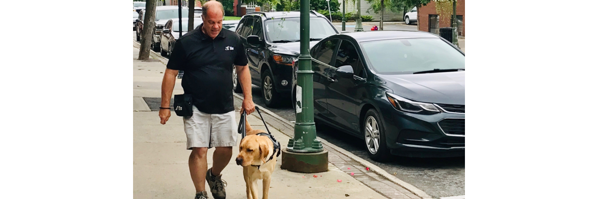 Carl marchant sur un trottoir avec un chien-guide d'INCA en formation.]