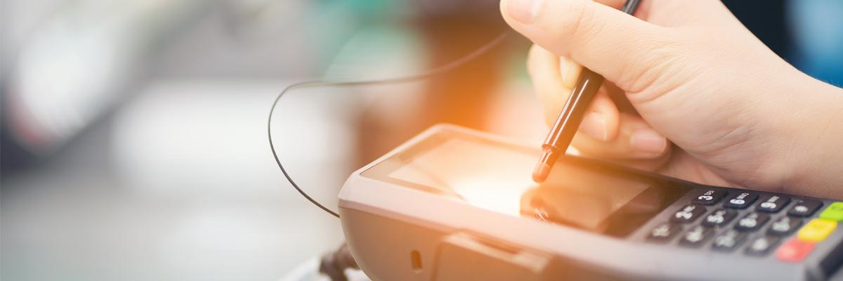 A hand using a stylus pen to make a selection on the touch screen surface of a payment terminal.